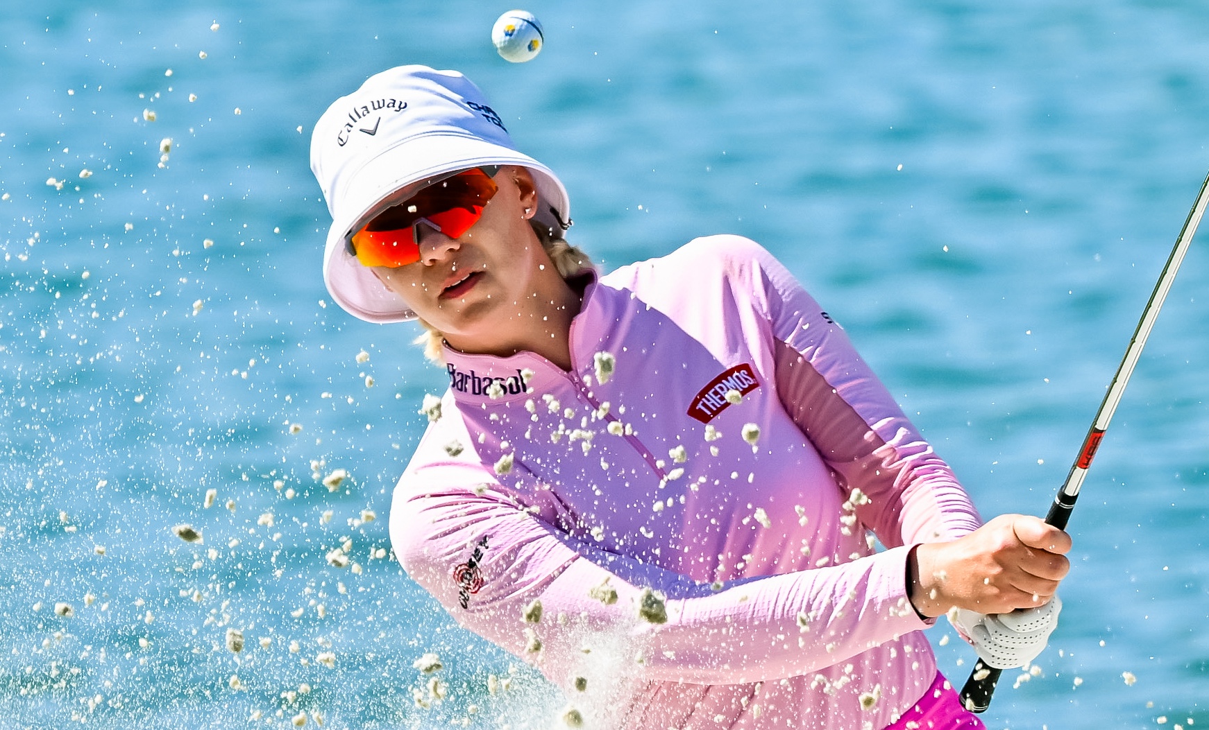 Madelene Sagstrom follows through on a bunker shot. The ball is popped up in the air and sand splashes up from the impact.