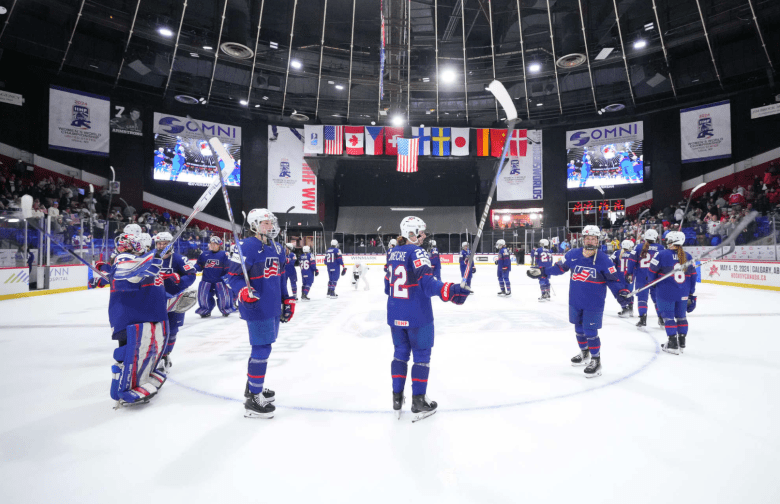 U.S. players raise their sticks in celebration at center ice. They are wearing blue uniforms.