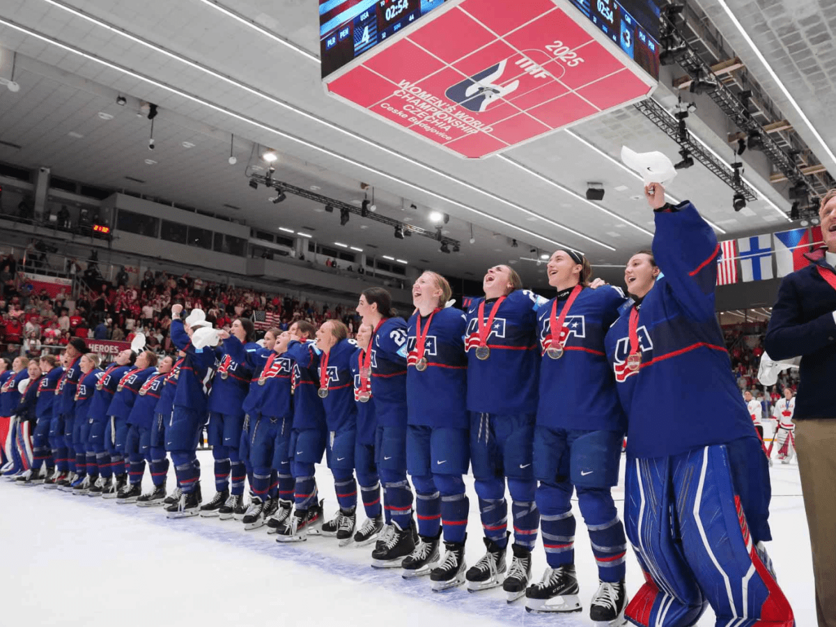 Team USA stands arm in arm on the blueline, belting along with their anthem as it plays on the speakers. They are wearing blue uniforms.
