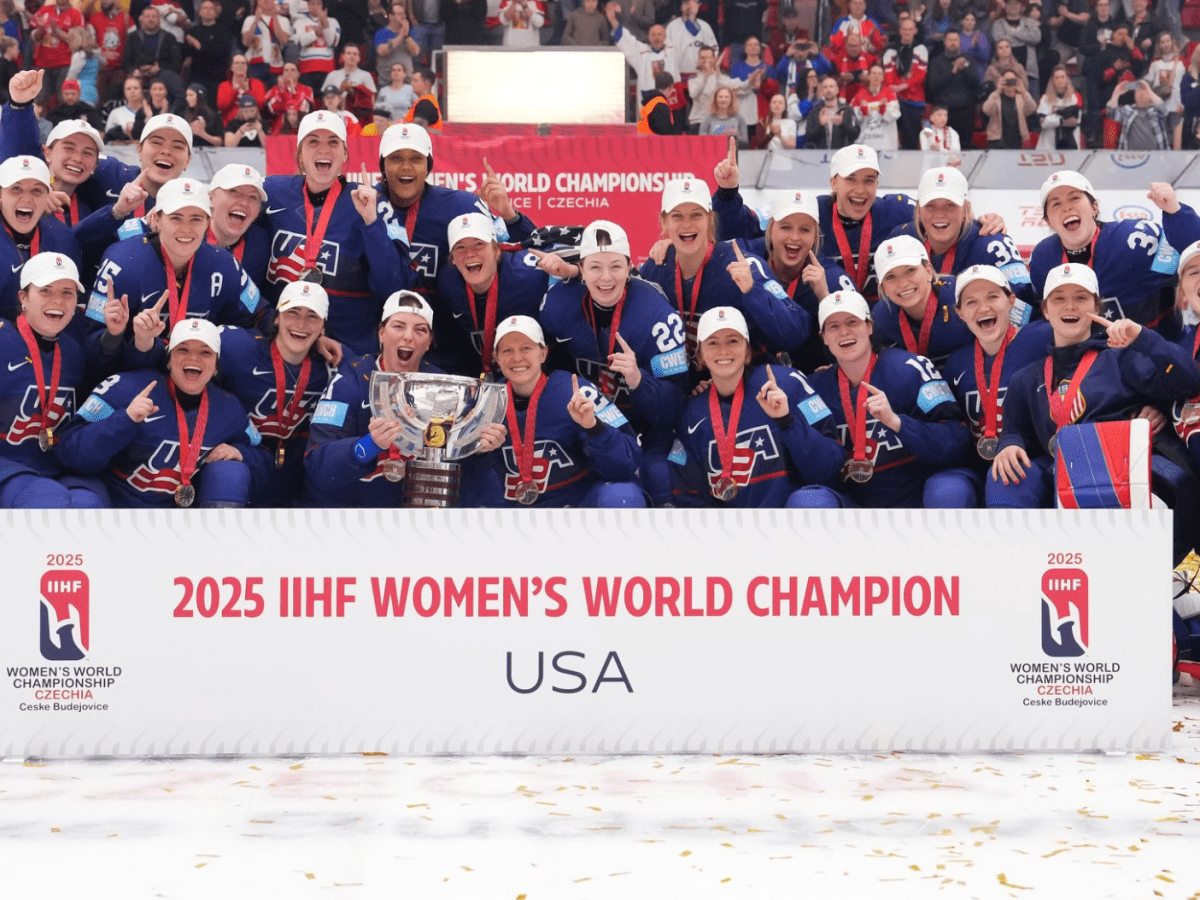 Team USA crowds together behind a sign that says "2025 IIHF Women's World Champion USA. They all smile while holding up one finger.