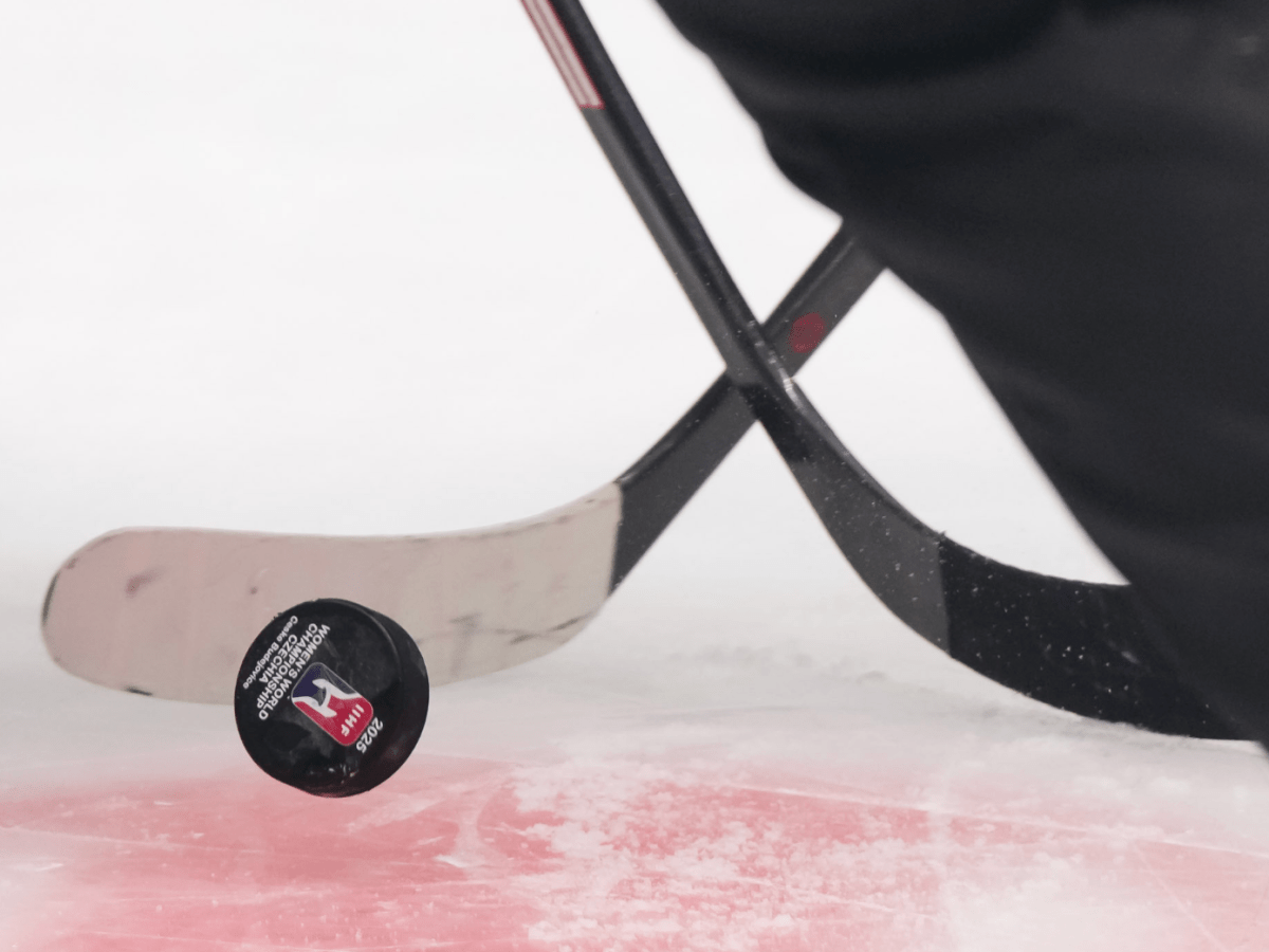 An IIHF branded puck has just landed on the ice, as two cross sticks begin to battle for it.