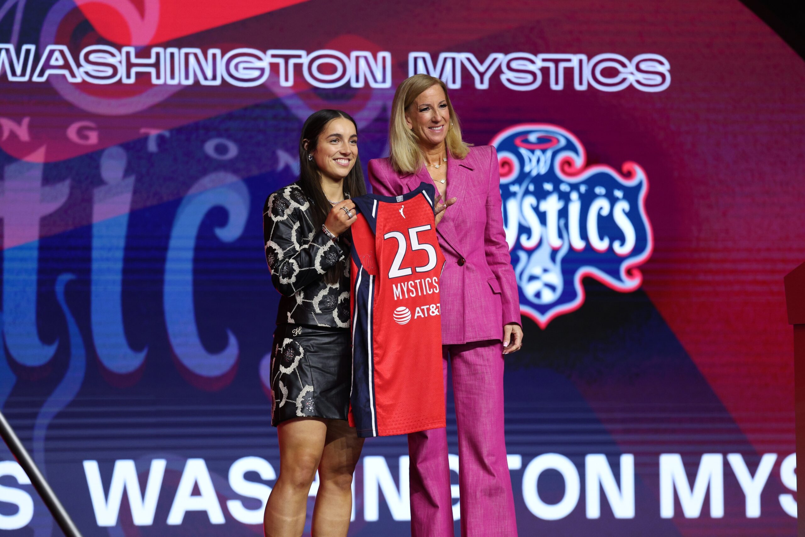 Kentucky guard Georgia Amoore and WNBA Commissioner Cathy Engelbert hold up a red Washington Mystics No. 25 jersey. They smile for a photo in front of a backdrop showing the Mystics' name and logo.