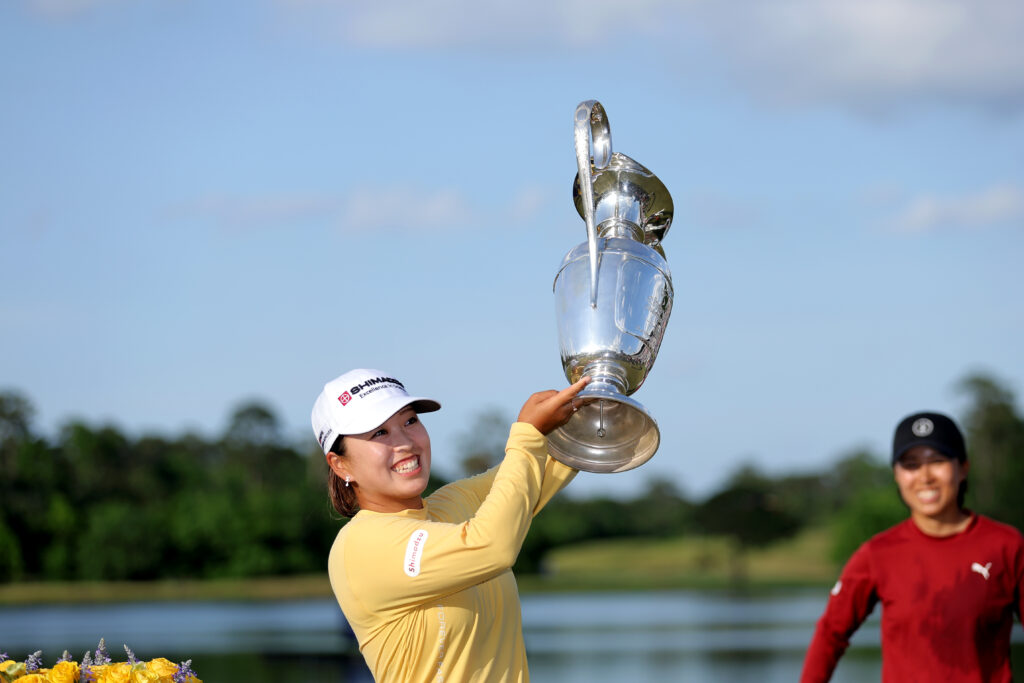 Mao Saigo holds up The Chevron Championship trophy high to her left and smiles widely. A friend supports her in the background.