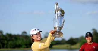 Mao Saigo holds up The Chevron Championship trophy high to her left and smiles widely. A friend supports her in the background.