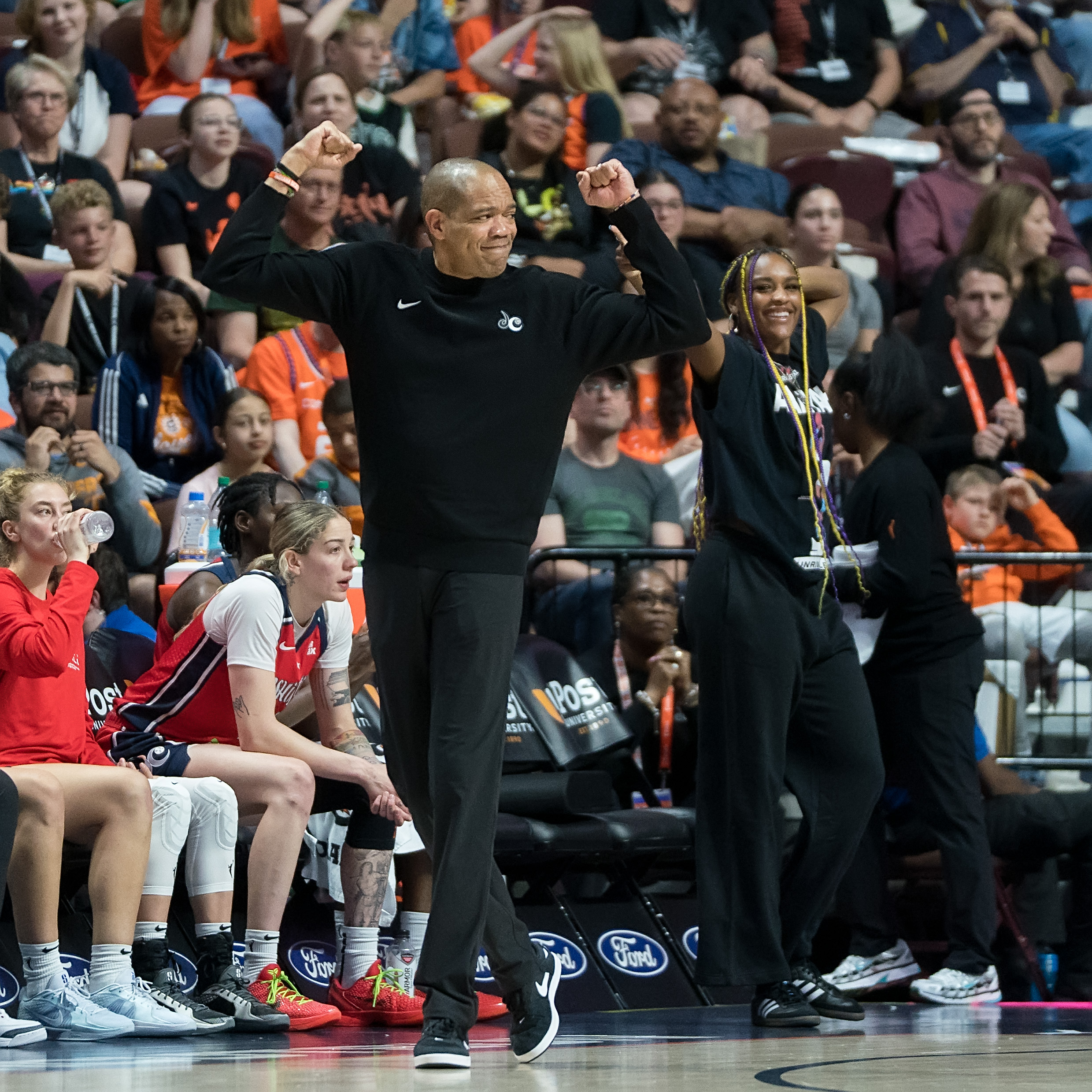 Washington Mystics head coach Sydney Johnson raises both arms and pumps his fists in celebration. Forward Aaliyah Edwards stands on the sideline behind him and makes the arm gestures that referees use to call a charge.