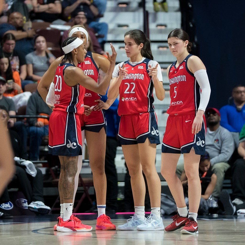 Washington Mystics guard Brittney Sykes gestures with her right hand to wing Sonia Citron in a huddle. Citron points with both hands toward the court. Forward Kiki Iriafen is behind them listening with her hands on her hips, and guard Jade Melbourne stands a step away from the huddle, looking out at the court.