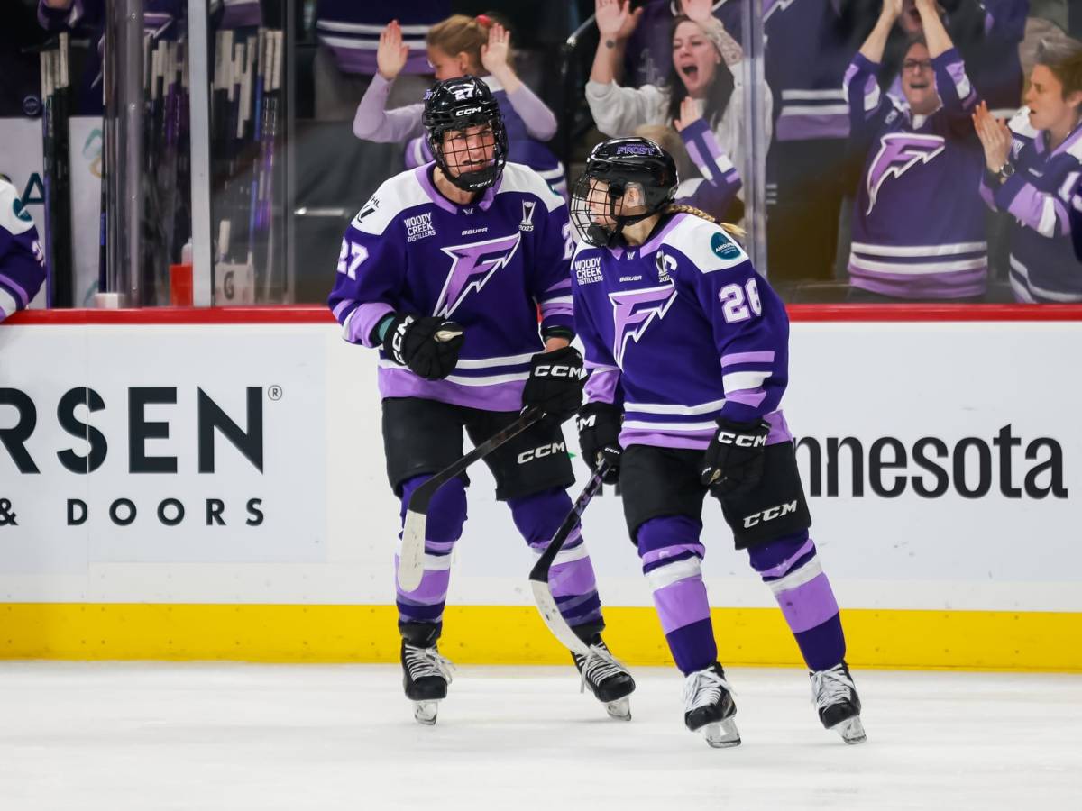 Heise (left) skates over to Coyne Schofield (right) as fans cheer in celebration behind them. They are both wearing purple home uniforms.