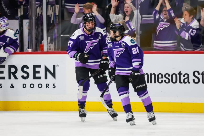 Heise (left) skates over to Coyne Schofield (right) as fans cheer in celebration behind them. They are both wearing purple home uniforms.
