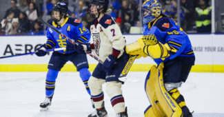 During a PWHL game between Toronto and Montréal, Laura Stacey stands in front of Kristen Campbell, who is trying to look around her. Renata Fast is coming in to defend. Stacey is wearing a cream away uniform, while Campbell and Fast are in blue.
