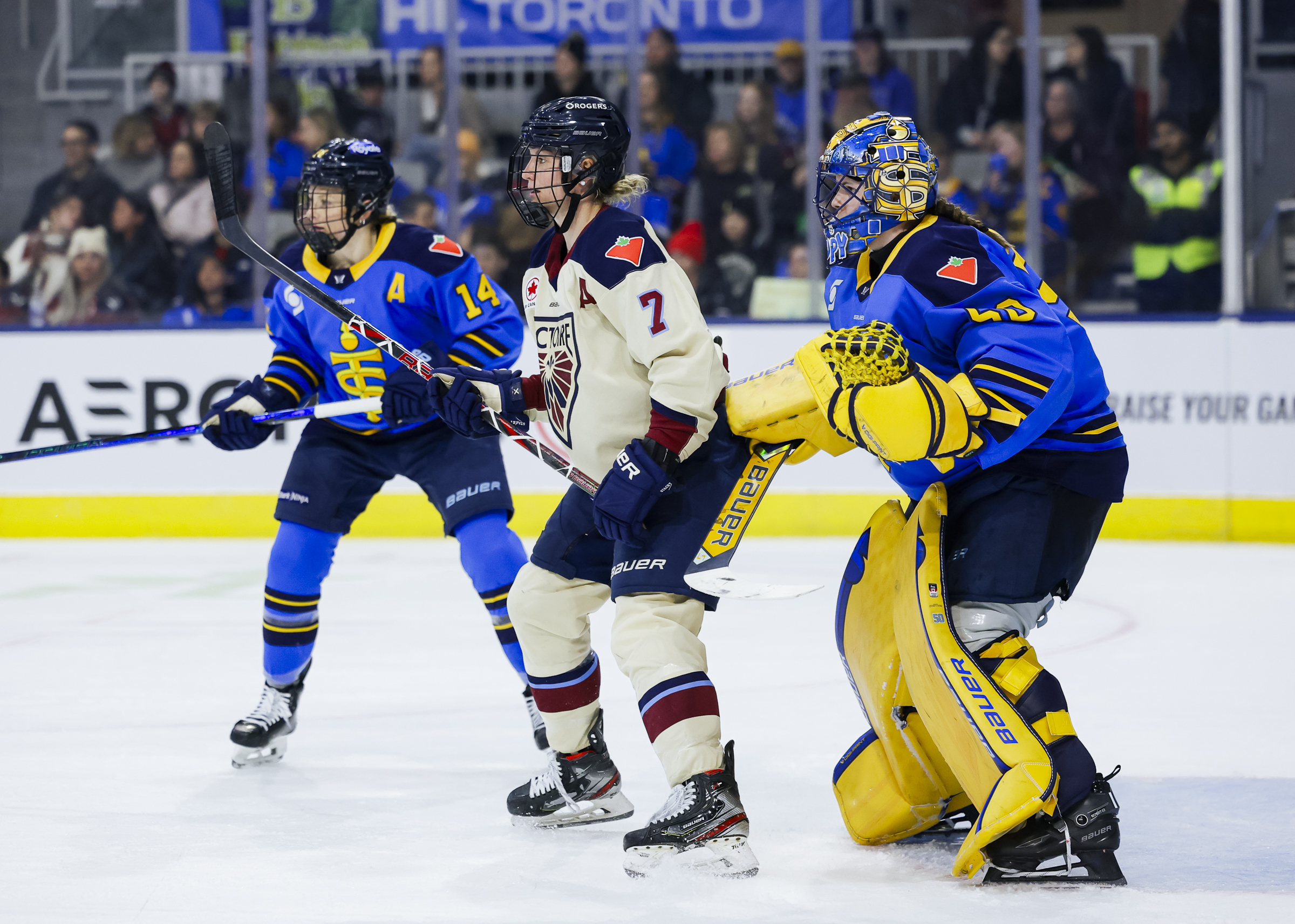 During a PWHL game between Toronto and Montréal, Laura Stacey stands in front of Kristen Campbell, who is trying to look around her. Renata Fast is coming in to defend. Stacey is wearing a cream away uniform, while Campbell and Fast are in blue.
