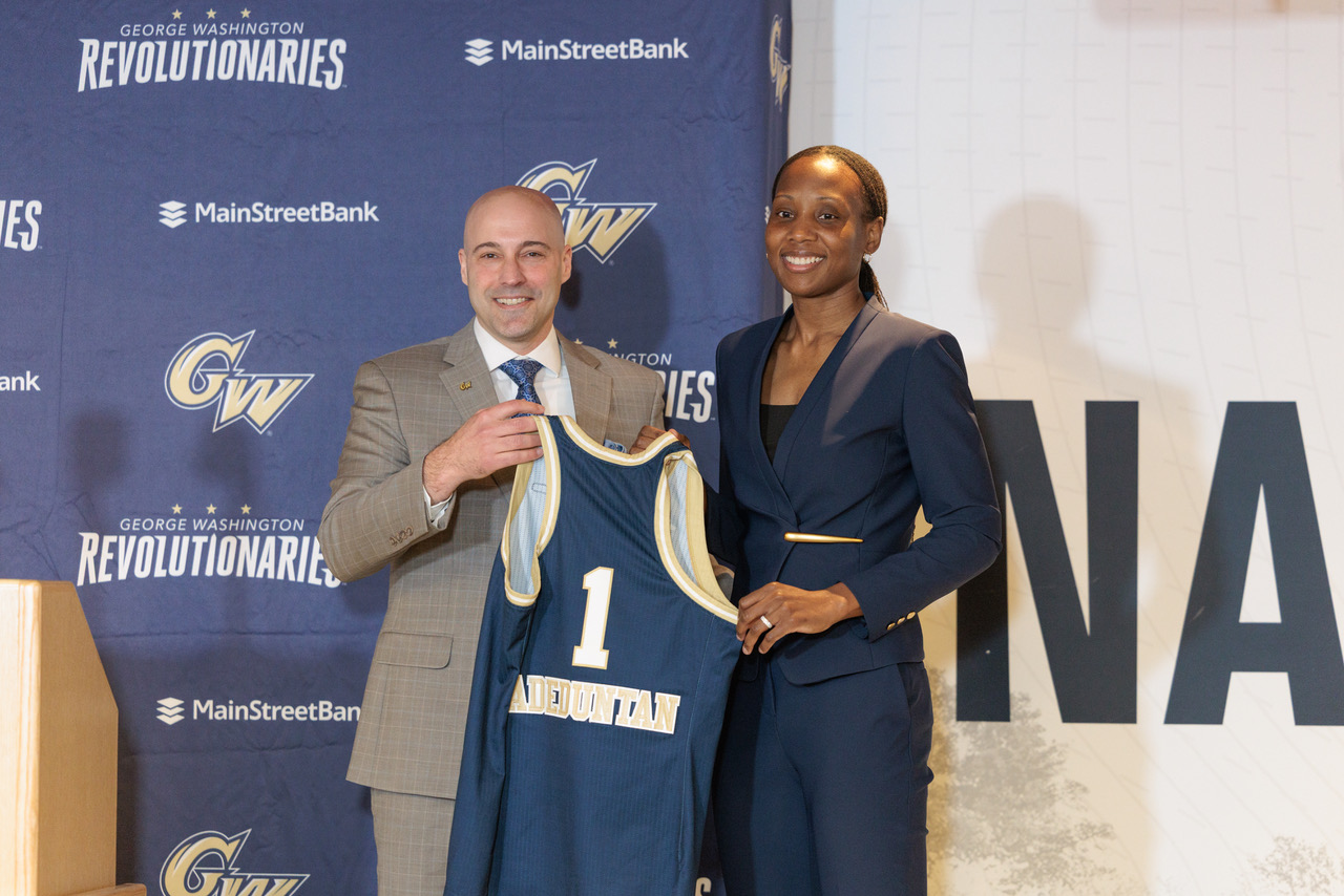 George Washington head coach Ganiyat Adeduntan (right) poses for a photo with associate vice president and director of athletics Michael Lipitz (left). They are both smiling and holding up a GW jersey with No. 1 and “Adeduntan” on the back.