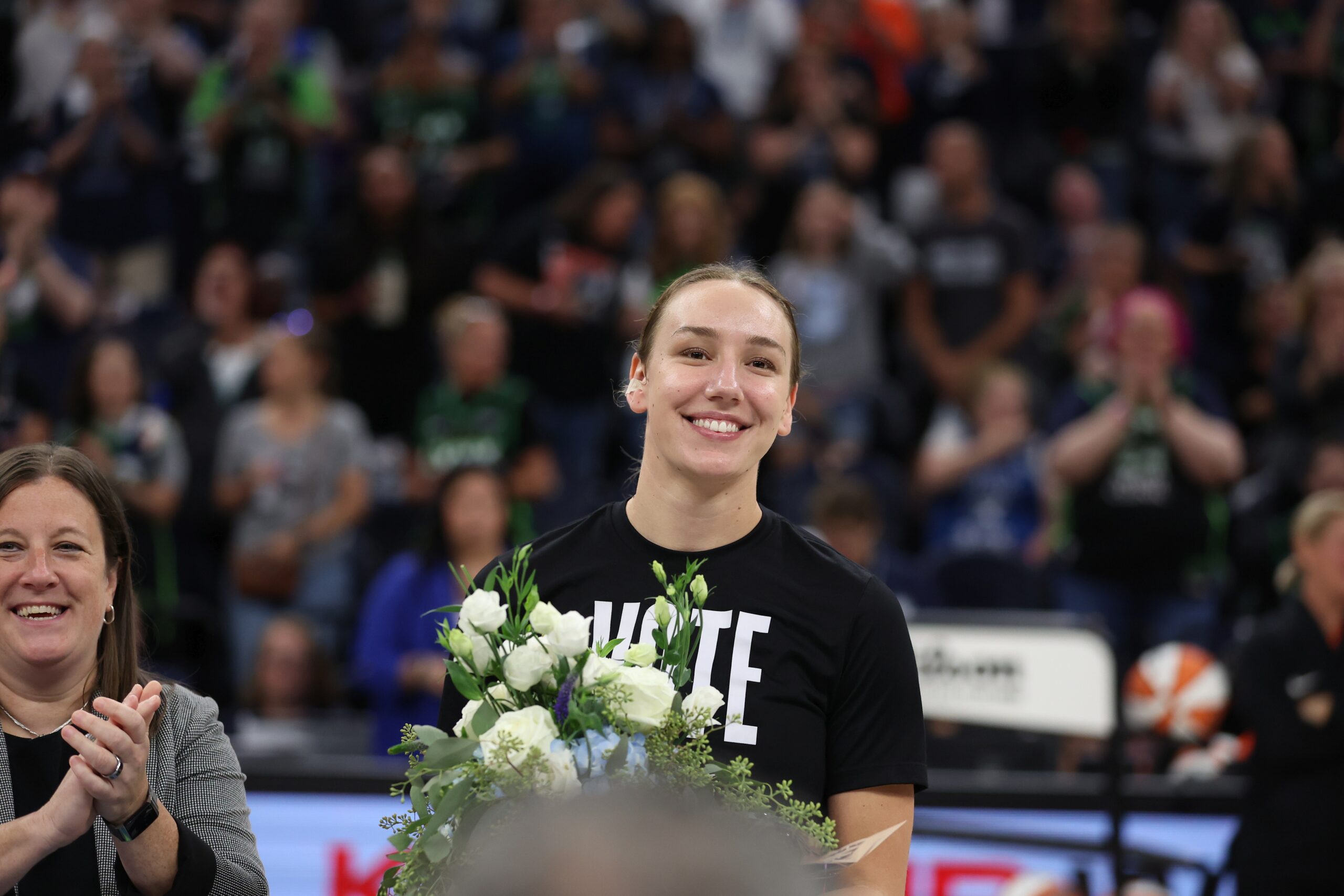 Alanna Smith smiles while holding flowers in front of a sold out arena.