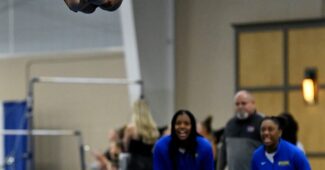 A gymnast in a blue and yellow leotard performs a piked flip in the air as her team watches below.