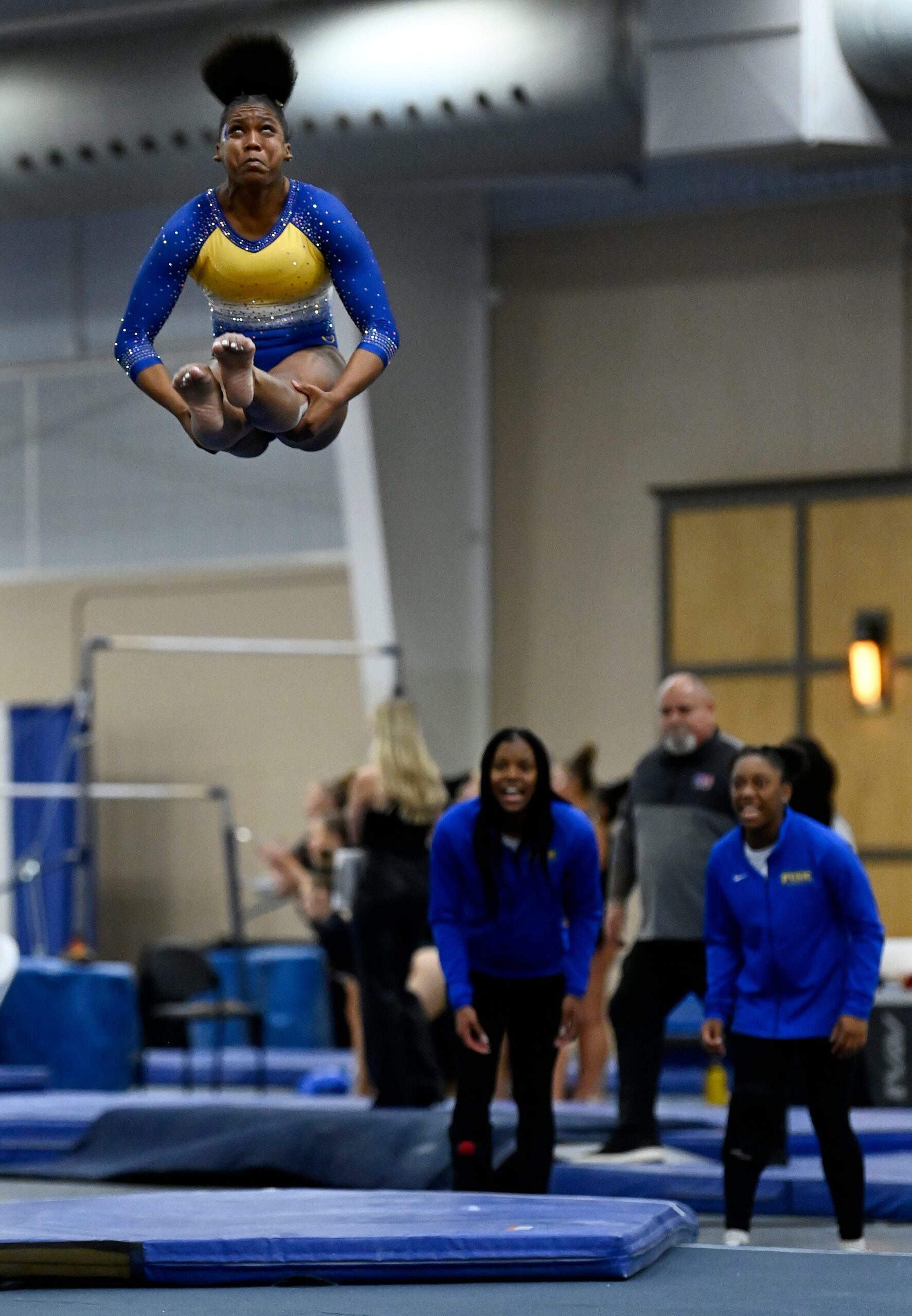 A gymnast in a blue and yellow leotard performs a piked flip in the air as her team watches below.