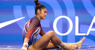 An athlete in a red, white, and blue leotard sits on a mat with a disgruntled expression on her face.