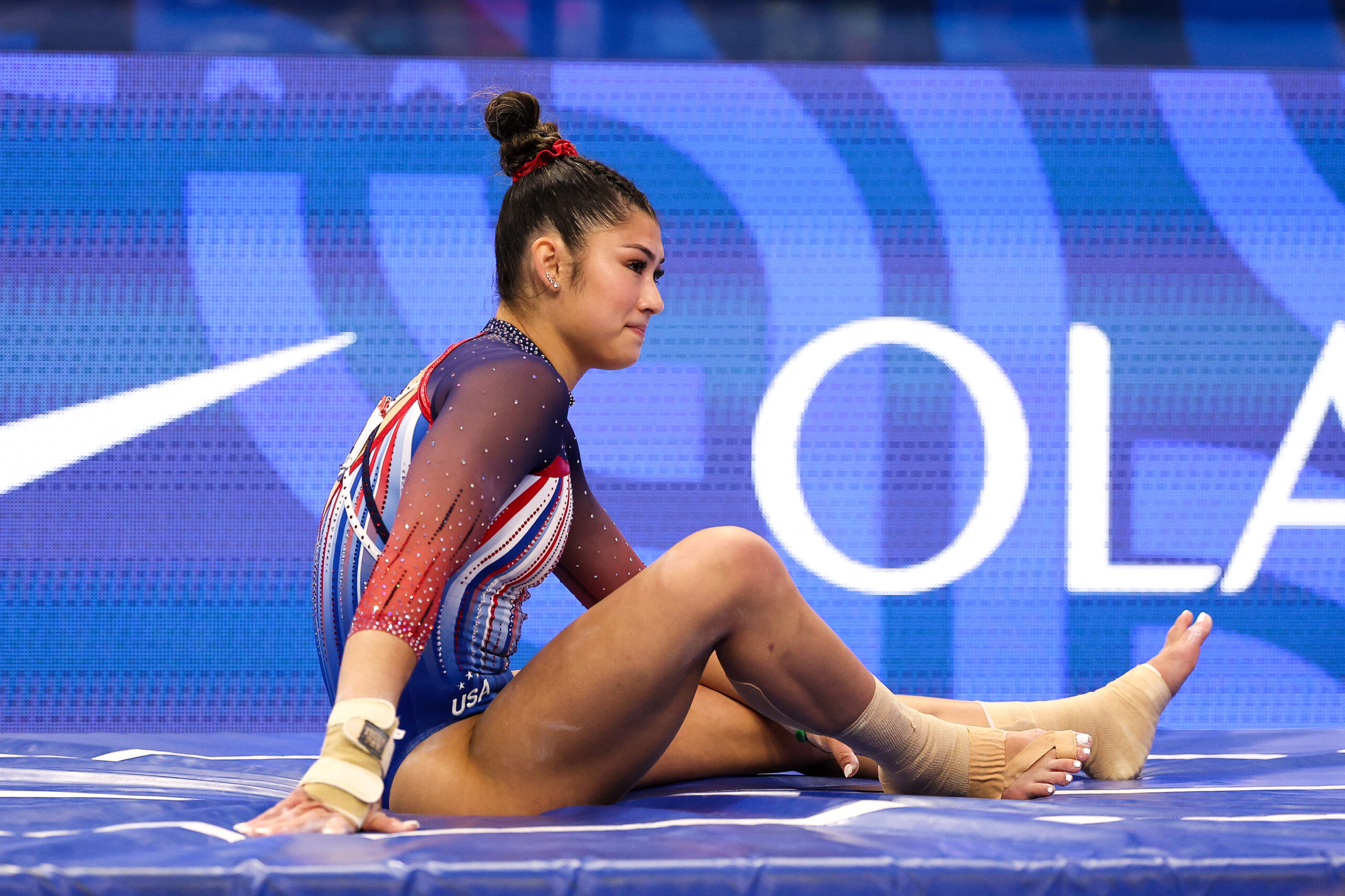 An athlete in a red, white, and blue leotard sits on a mat with a disgruntled expression on her face.
