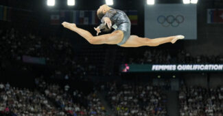 A gymnast in a black and silver leotard does a leap over the balance beam.