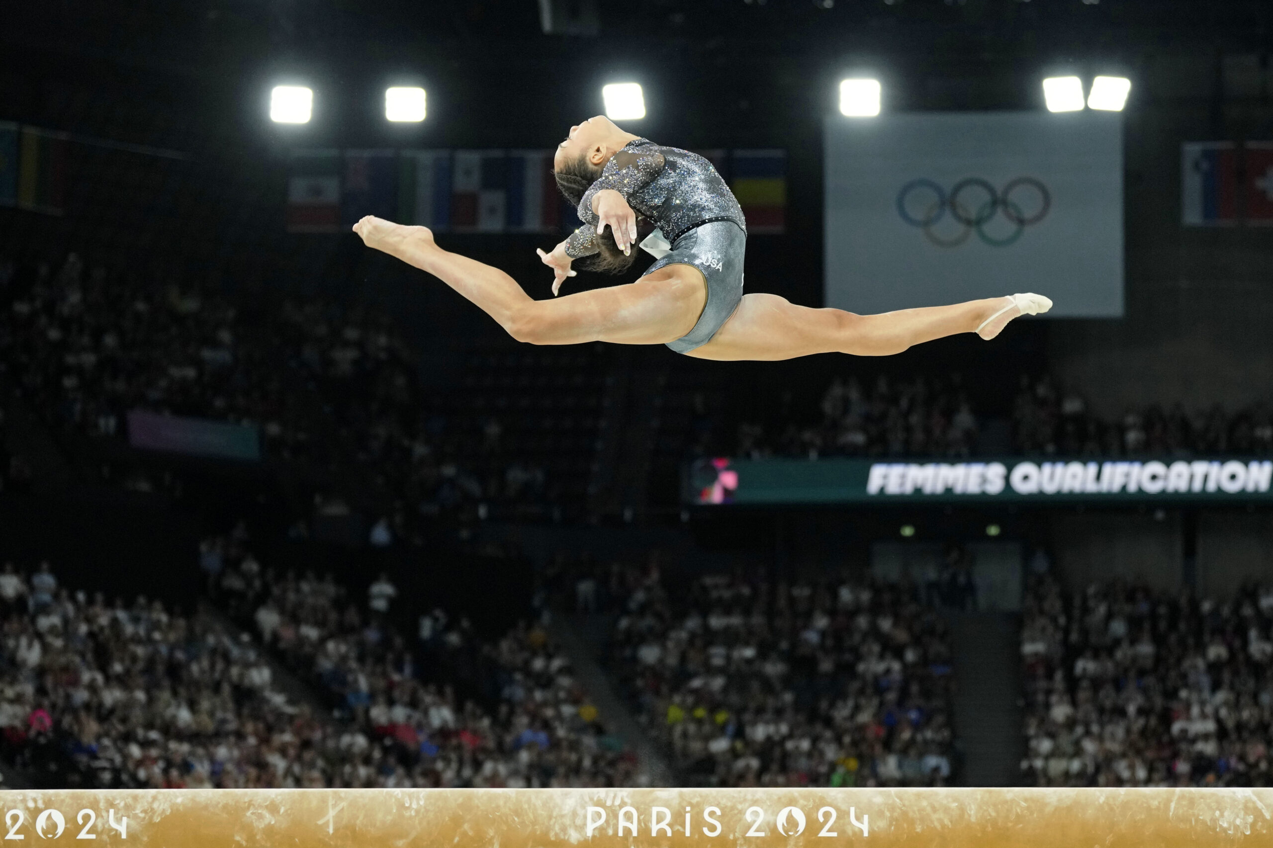 A gymnast in a black and silver leotard does a leap over the balance beam.
