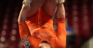 A gymnast in an orange leotard does a flip in the air in a pike position.