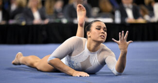 A gymnast in a white leotard holds up her hand while competing on floor exercise.