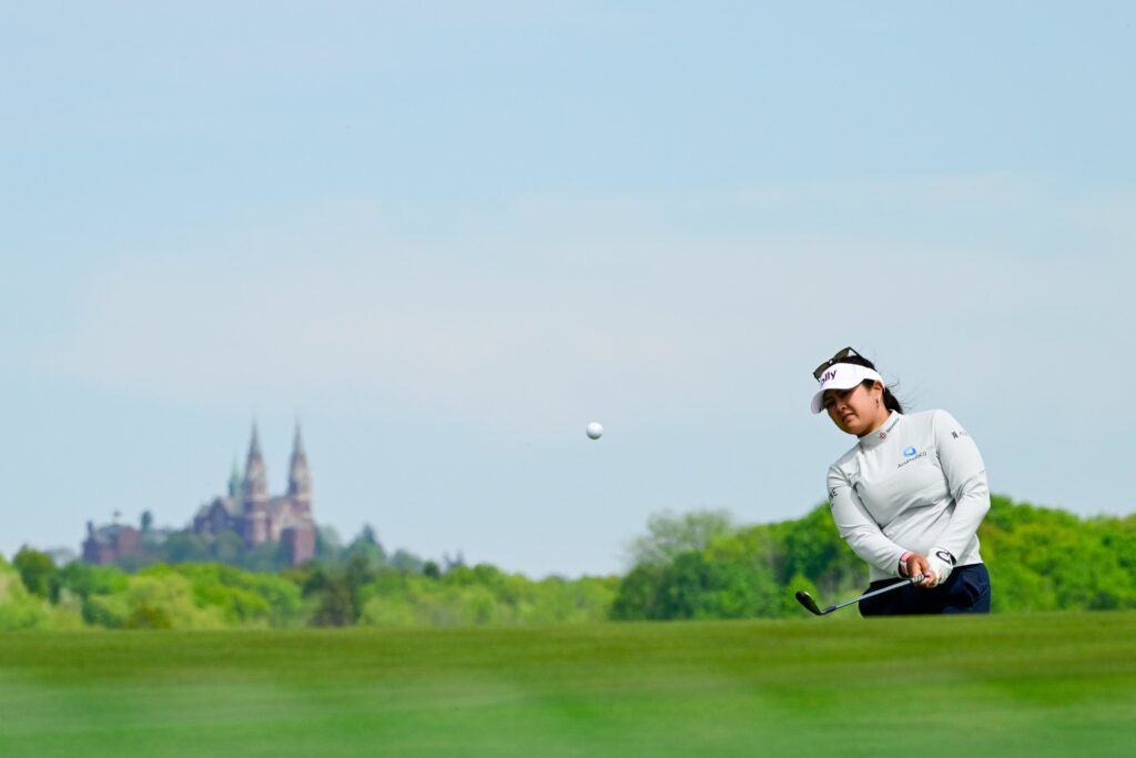 Lilia Vu is on the right side of the picture chipping a shot onto the green toward the camera. In the background is the vast landscape of Erin Hills with a looming building.