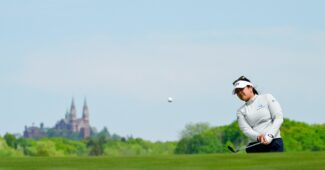 Lilia Vu is on the right side of the picture chipping a shot onto the green toward the camera. In the background is the vast landscape of Erin Hills with a looming building.