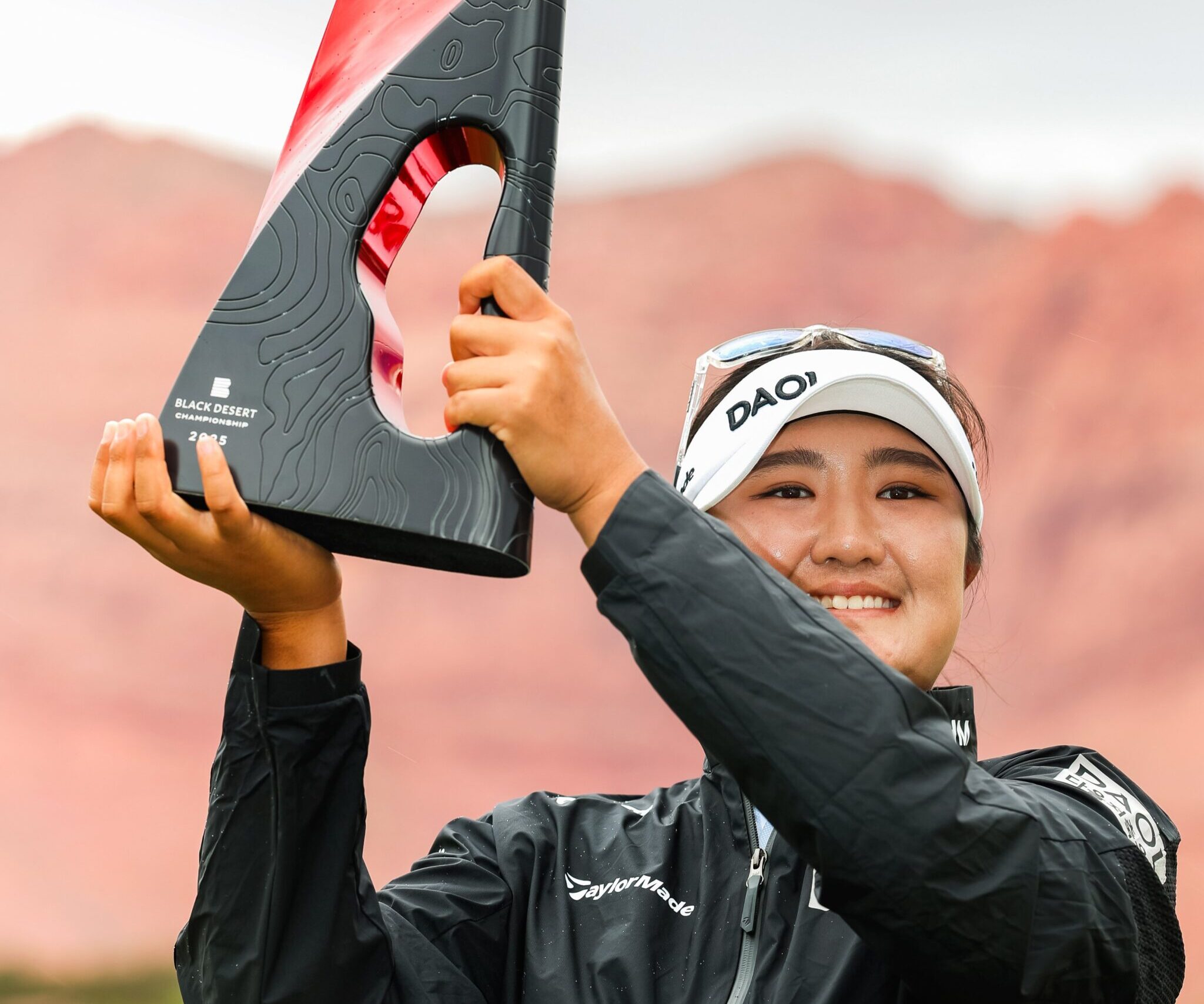 Haeran Ryu holds up a shiny red and textured black Black Desert Championship trophy to her right. She smiles and looks beyond the camera.