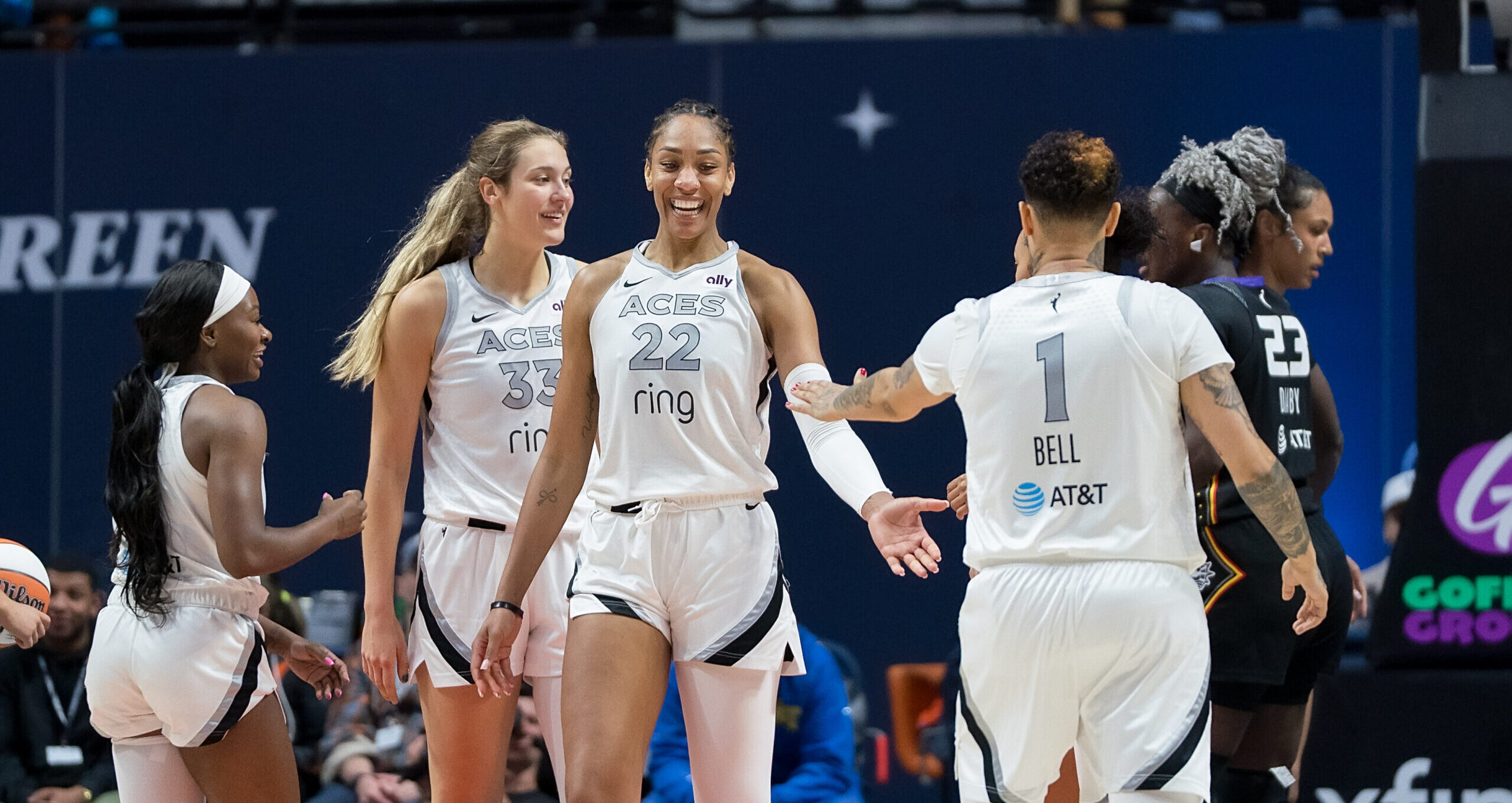 Las Vegas Aces forward Kierstan Bell faces away from the camera and reaches out to dap up teammate A'ja Wilson who is standing and facing the camera with a big smile as other teammates talk and smile behind them.