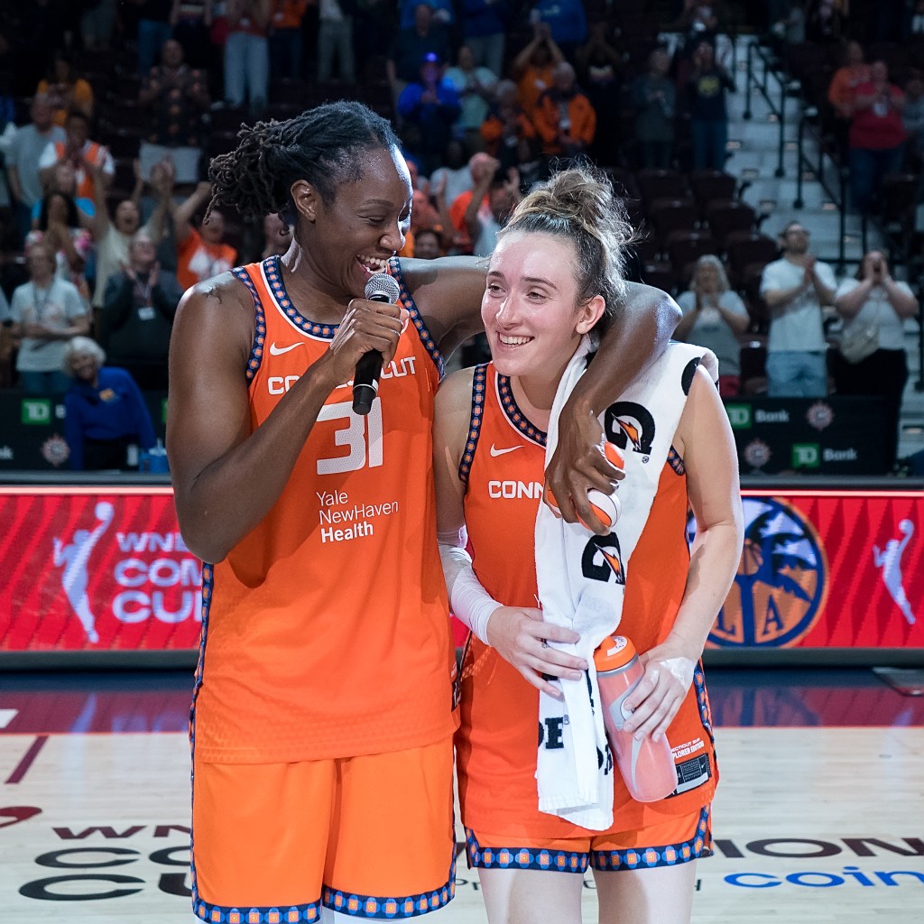 Connecticut Sun center Tina Charles speaks into a microphone after a win. She has her left arm draped around the shoulders of guard Marina Mabrey, and both players are smiling.
