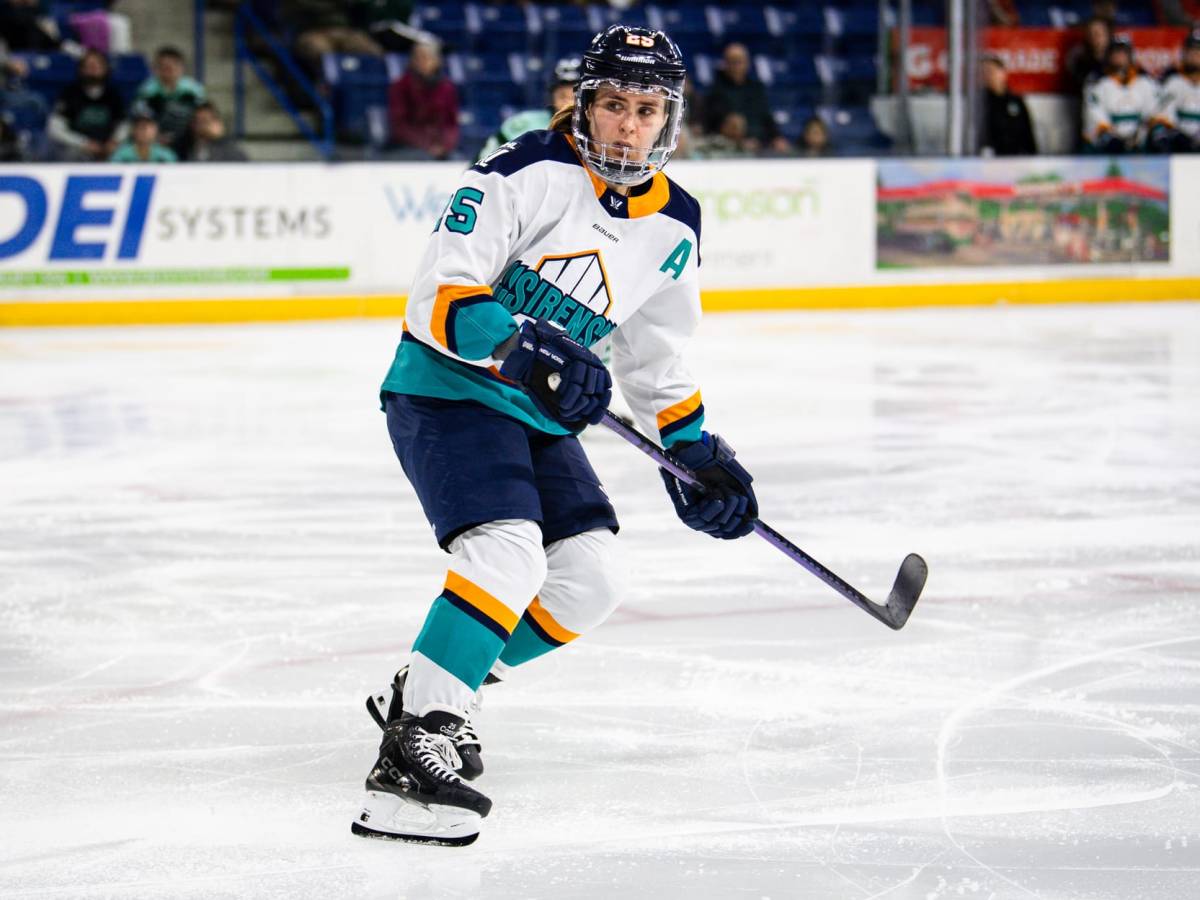 Carpenter is beginning to skate up ice as she looks behind her, watching the play unfold. She is wearing a white away uniform.