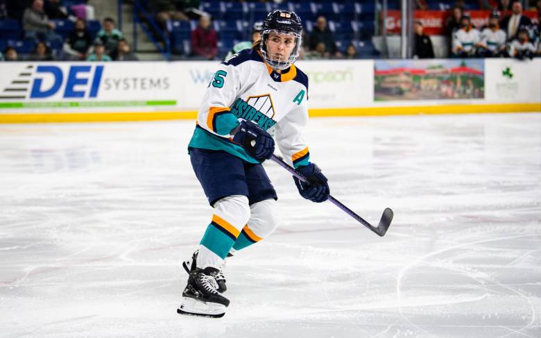 Carpenter is beginning to skate up ice as she looks behind her, watching the play unfold. She is wearing a white away uniform.