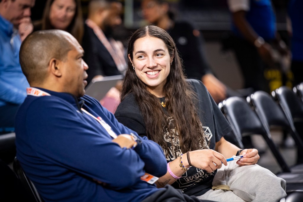Washington Mystics guard Georgia Amoore and head coach Sydney Johnson sit next to each other on the sidelines before a game. Johnson talks with his arms folded loosely across his chest, and Amoore smiles and looks at him as he talks.