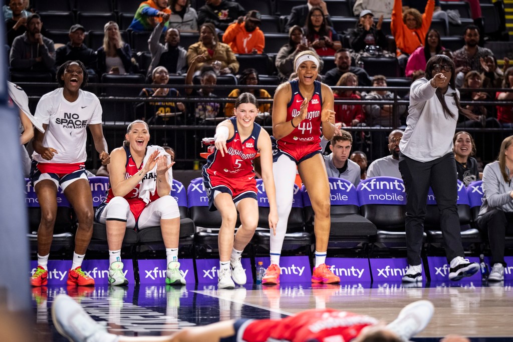 Washington Mystics guard Jade Melbourne, forward Kiki Iriafen and assistant coach Barbara Turner all stand up on the sideline to celebrate a play. Iriafen claps, Turner points to something on the court with her right hand, and Melbourne has her right arm extended with two fingers pointing down.