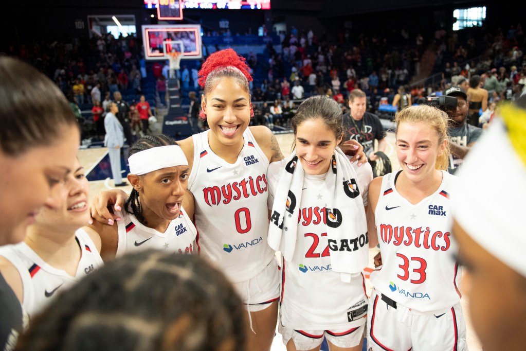 The Washington Mystics huddle at center court after a game. Center/forward Shakira Austin smiles widely and sticks her tongue out slightly as guard Brittney Sykes talks.