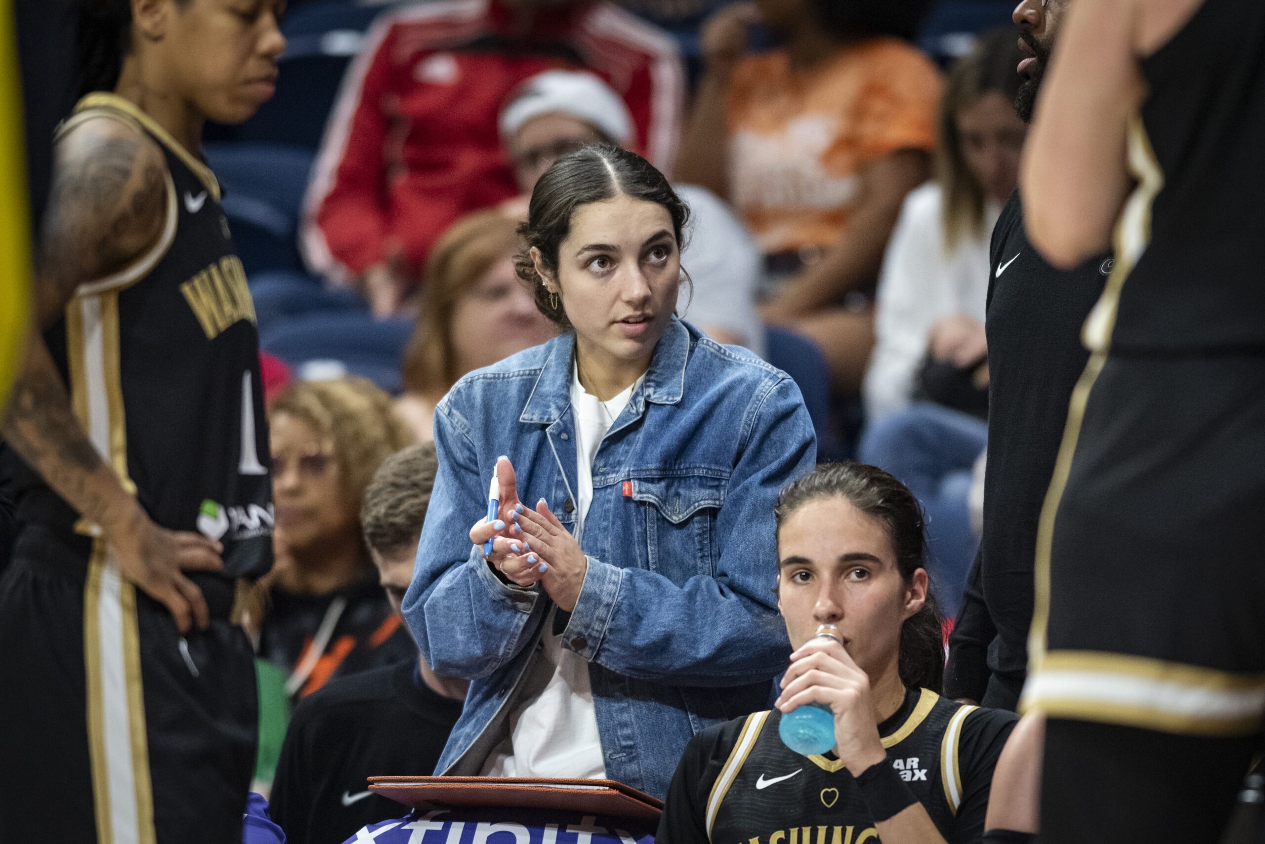 Washington Mystics guard Georgia Amoore stands in the second row on the Mystics' sideline and claps her hands together. Wing Sonia Citron sits in front of her and sips on a blue recovery drink.