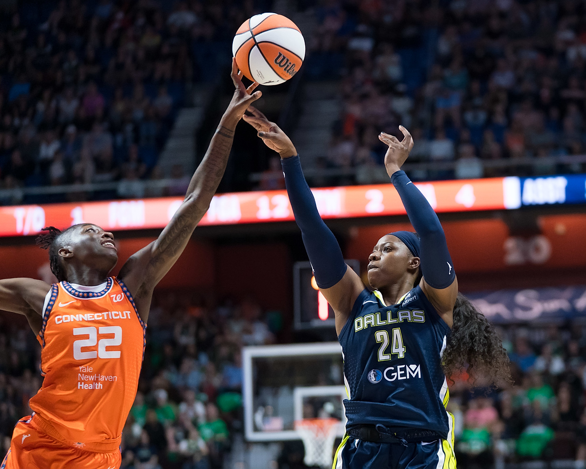 Connecticut Sun wing Saniya Rivers blocks a jump shot by Dallas Wings guard Arike Ogunbowale using her outstretched fingertips. A packed arena is out of focus in the background.