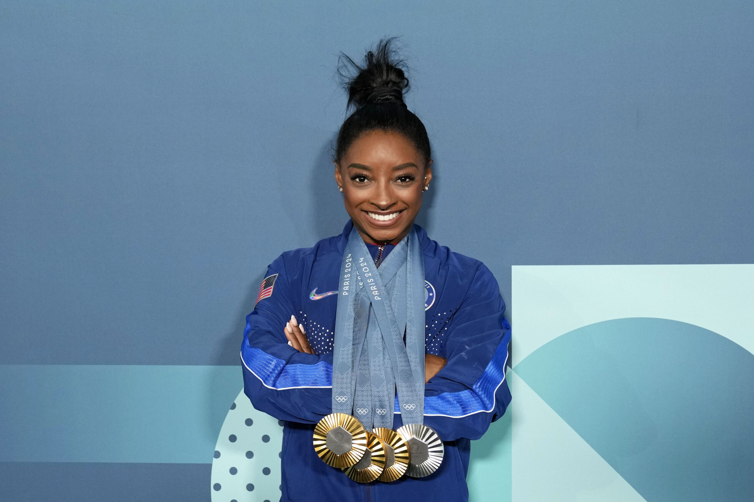Simone Biles in a blue tracksuit posing with all of her Olympic medals in Paris last summer.