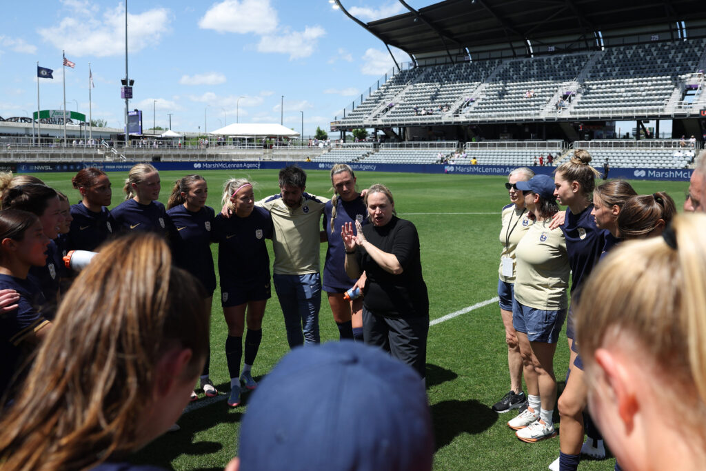 A group of players and coaches stand in a huddle on a soccer field