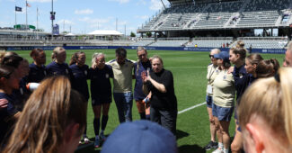 A group of players and coaches stand in a huddle on a soccer field