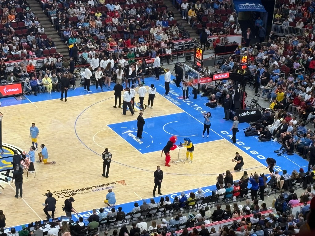 In the lower right corner of a basketball court, viewed from above, a bull mascot and a lion mascot play Connect Four at a small bar-height table.