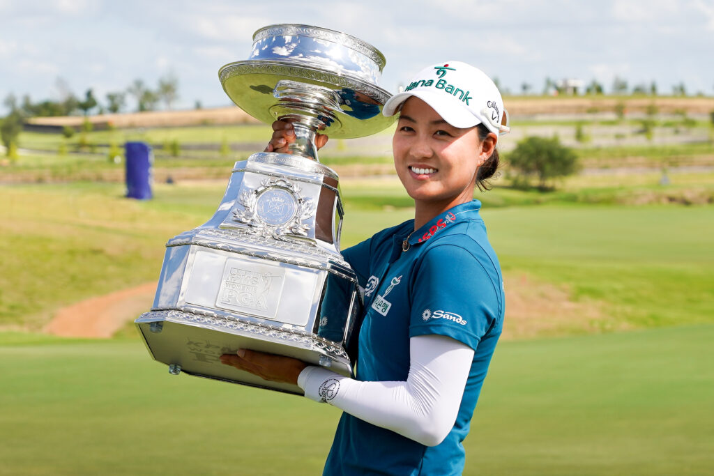 Minjee Lee holds up the KPMG Women's PGA Championship to her left and smiles for a picture. The golf course Fields Ranch East stretches out behind her.