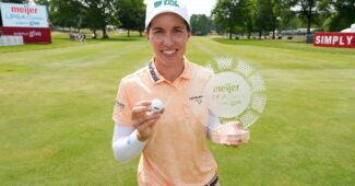 Carlota Ciganda holds up the Meijer LPGA Classic trophy in her left hand and her winning golf ball in her right hand, smiling. The fairway of the golf course with leaderboard and Simply Give signage stretches behind her.