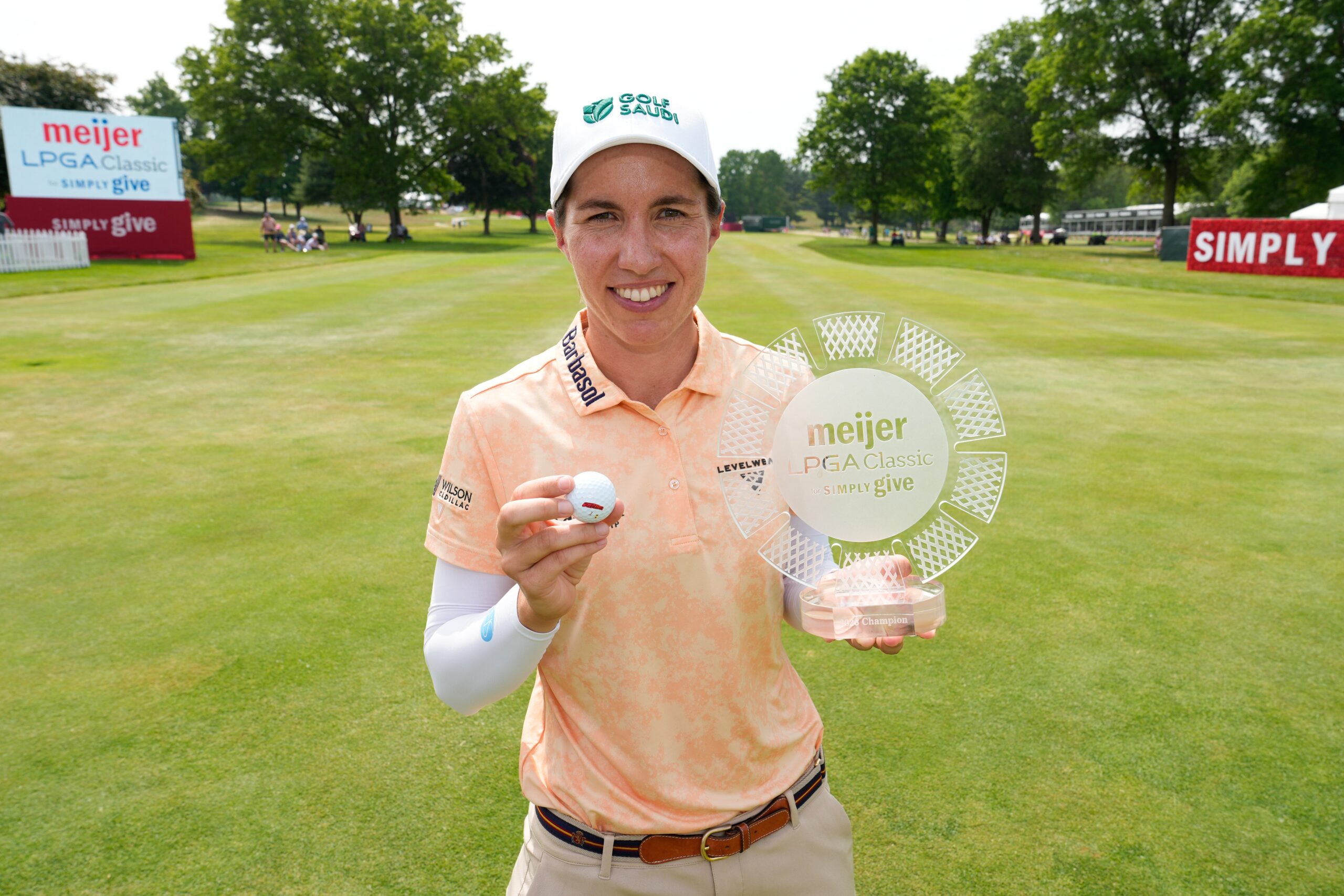 Carlota Ciganda holds up the Meijer LPGA Classic trophy in her left hand and her winning golf ball in her right hand, smiling. The fairway of the golf course with leaderboard and Simply Give signage stretches behind her.