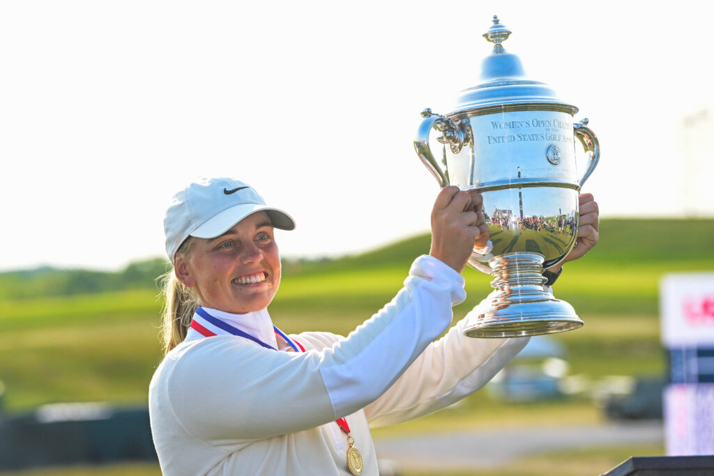 Maja Stark raises the U.S. Women's Open trophy to her left and smiles beyond the camera. The hills of Erin Hills golf course are in the background and the sun sets.