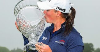 Jennifer Kupcho holds the 2025 ShopRite LPGA Classic trophy up to her right and kisses the crystal side. The sky is cloudy and overcast.