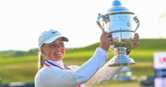 Maja Stark raises the U.S. Women's Open trophy to her left and smiles beyond the camera. The hills of Erin Hills golf course are in the background and the sun sets.