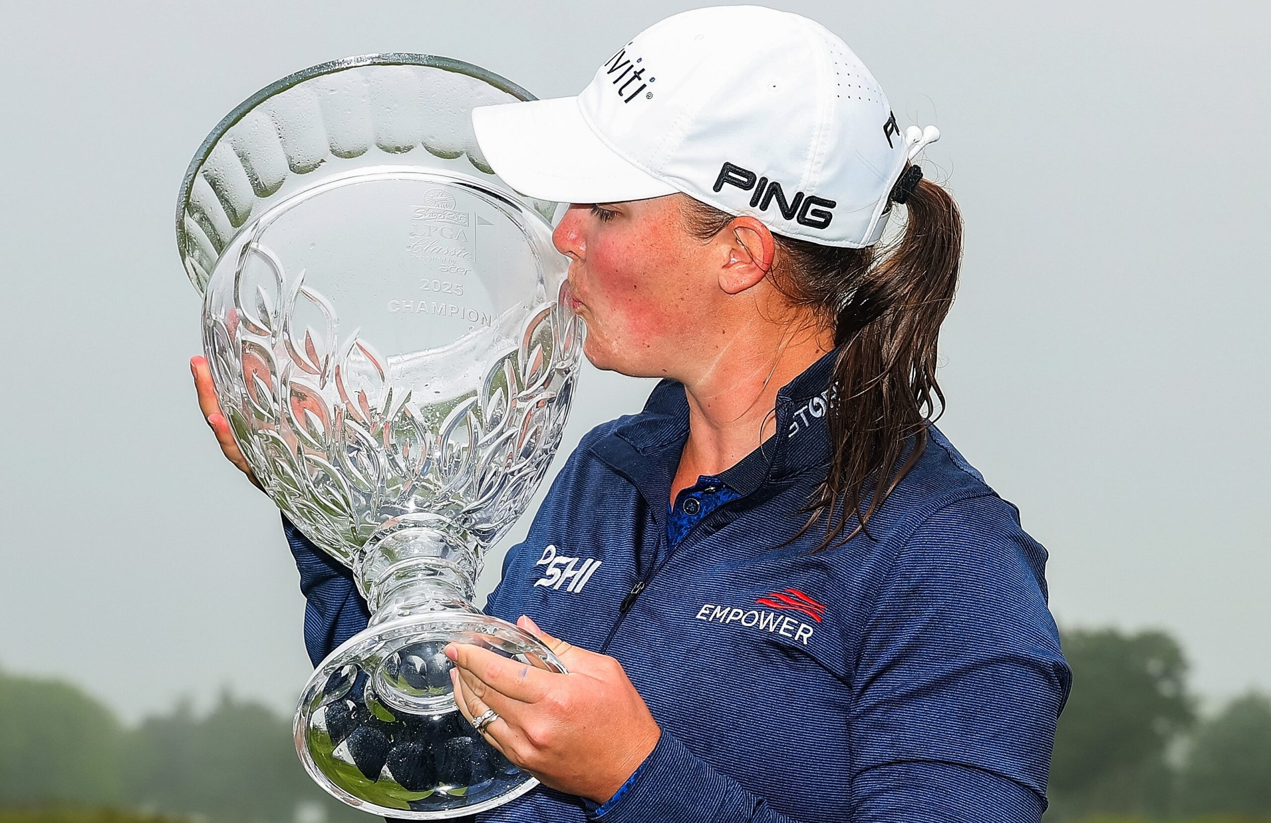 Jennifer Kupcho holds the 2025 ShopRite LPGA Classic trophy up to her right and kisses the crystal side. The sky is cloudy and overcast.
