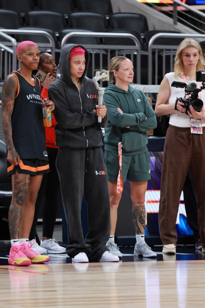 Courtney Williams and Natisha Hiedeman stand on the sideline with a tripod and a camera in Hiedeman's hand.