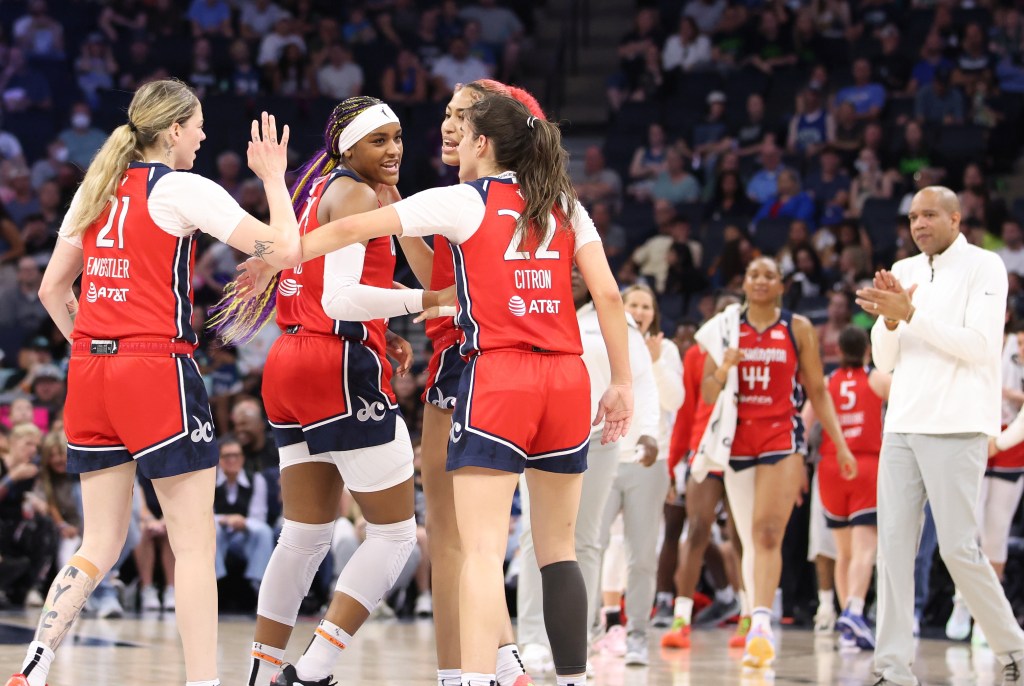 Four Washington Mystics players approach each other during a stoppage in play. A few have arms out for high-fives. Head coach Sydney Johnson is visible in the background, approaching the group and clapping.
