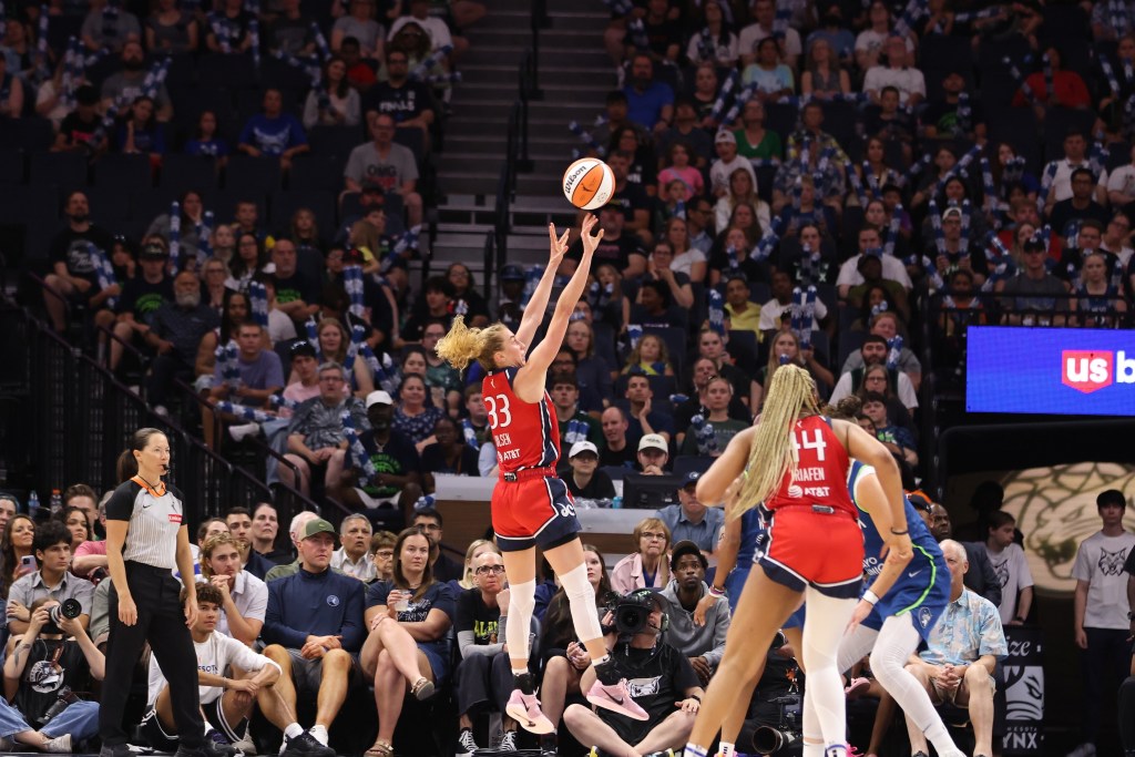 Washington Mystics guard Lucy Olsen is shown from the side, shooting an open midrange jump shot. The ball has just come off her fingertips, and no one has a hand up to contest the shot.